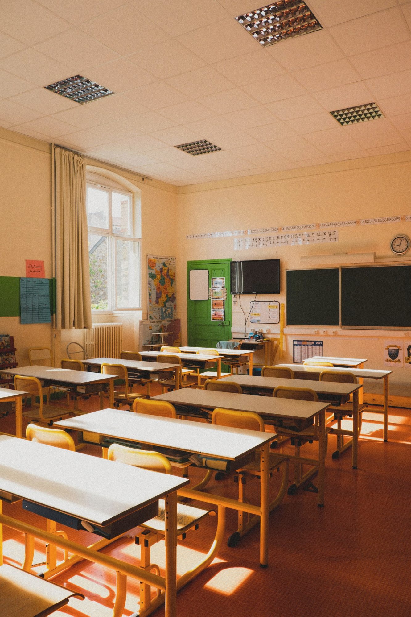 A sunlit primary classroom with rows of desks and yellow chairs, before the students arrive